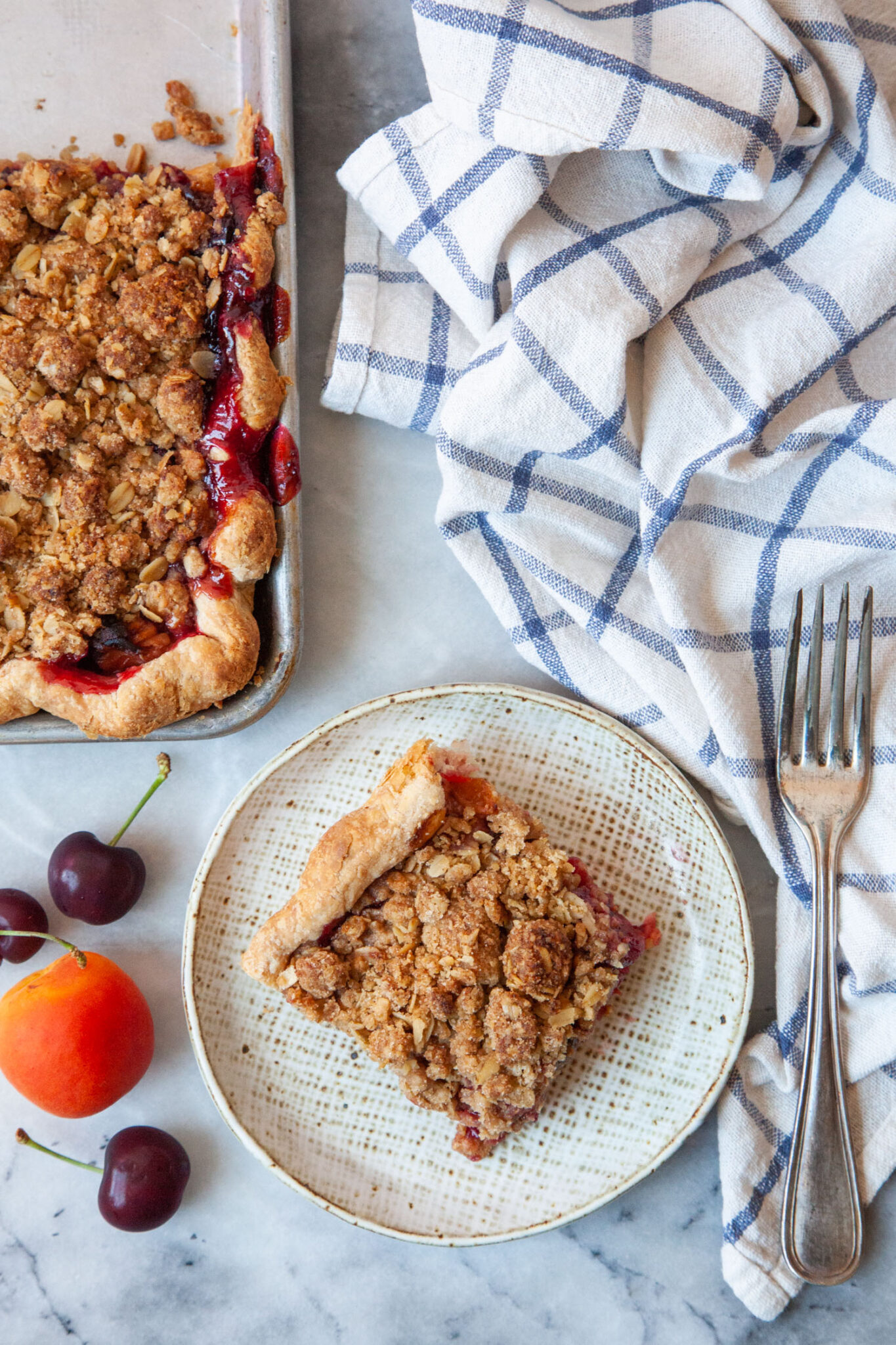 A slice of apricot cherry slab pie on a plate, with the remaining slab pie next to it. There are fresh cherries and an apricot next tot he plate, along with a blue lined cloth napkin.